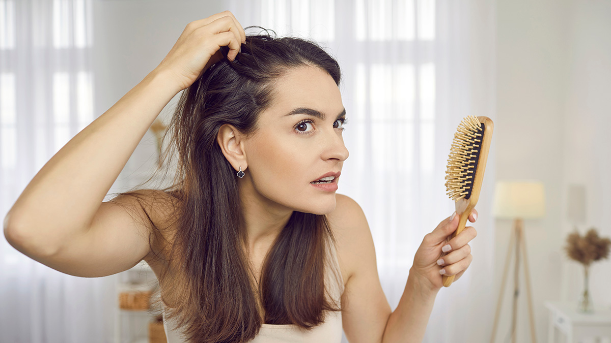 A young woman inspecting her scalp and worried about hair loss while holding a brush in her other hand